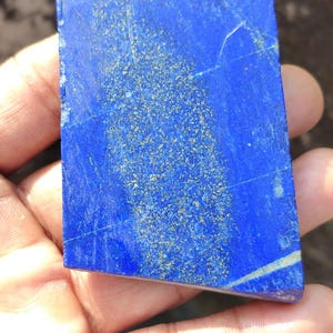 Può includere: Una lastra rettangolare di pietra lapislazzuli blu intenso caratterizzata da una prominente fascia centrale di inclusioni di pirite color oro scintillante e sottili venature di calcite bianca. La pietra ha una superficie lucida ed è contrassegnata dalla lettera D.