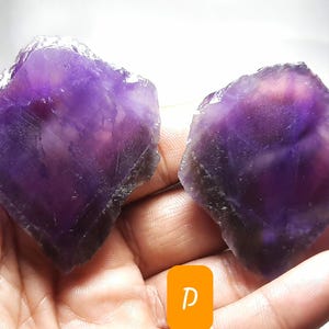 K&ouml;nnte beinhalten: Zwei rohe Amethystkristalle, jeder mit einem tiefvioletten Farbton und einer rauen, nat&uuml;rlichen Textur. Die Kristalle werden in einer Hand gehalten, wodurch ihre unregelm&auml;&szlig;igen Formen und das Lichtspiel auf ihren Oberfl&auml;chen zur Geltung kommen. Das Bild ist gut beleuchtet.