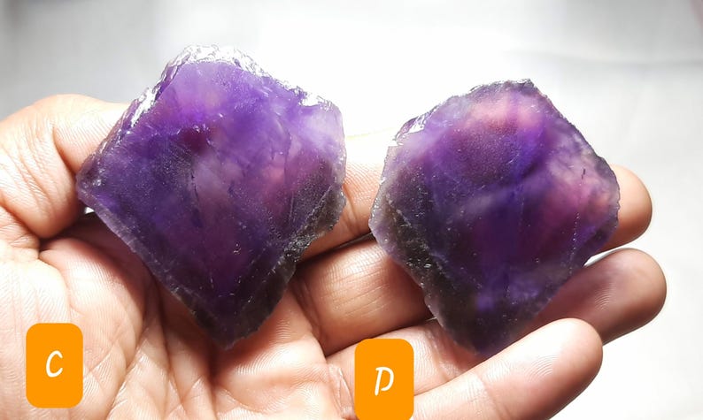 K&ouml;nnte beinhalten: Zwei rohe Amethystkristalle, jeder mit einem tiefvioletten Farbton und einer rauen, nat&uuml;rlichen Textur. Die Kristalle werden in einer Hand gehalten, wodurch ihre unregelm&auml;&szlig;igen Formen und das Lichtspiel auf ihren Oberfl&auml;chen zur Geltung kommen. Das Bild ist gut beleuchtet.