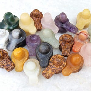 Könnte beinhalten: Eine Sammlung kleiner, geschnitzter Shiva-Lingam-Skulpturen aus Stein in verschiedenen Farben und Materialien, darunter Dunkelgrün, Weiß, Schwarz, Gelb, Braun, Rosa, Violett und Orange, angeordnet in einer Gruppe auf einer hellen, strukturierten Oberfläche.