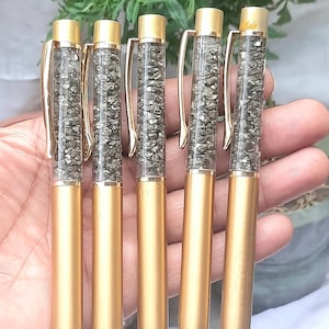 May include: Five gold-colored pens with clear barrels filled with small, irregularly shaped, gray stones. The pens have gold-colored clips and tips. The pens are held in a hand, with green foliage in the background.