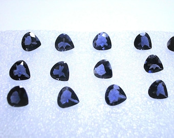 May include: A collection of thirteen loose, faceted gemstones with a deep blue color and a triangular shape. Each stone features multiple cut surfaces that reflect light, arranged in a grid pattern on a textured, white, foam-like surface.