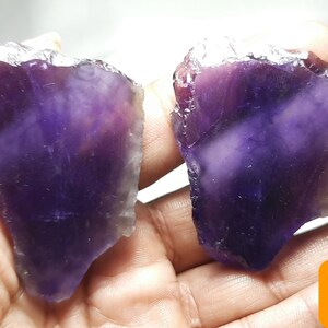 K&ouml;nnte beinhalten: Zwei rohe Amethyst-Kristallproben, jede mit einem tiefvioletten Farbton und einer rauen, nat&uuml;rlichen Textur. Die Kristalle werden in einer Hand gehalten, mit den Etiketten A und B in den Ecken. Die Amethyststeine haben eine durchscheinende Qualit&auml;t.