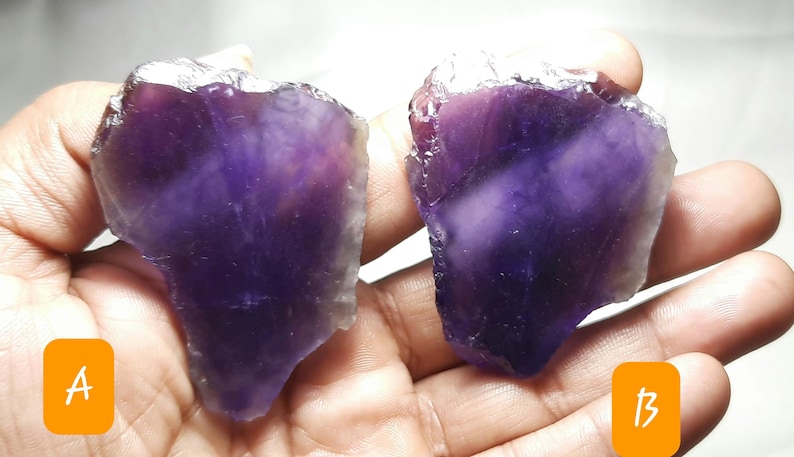K&ouml;nnte beinhalten: Zwei rohe Amethyst-Kristallproben, jede mit einem tiefvioletten Farbton und einer rauen, nat&uuml;rlichen Textur. Die Kristalle werden in einer Hand gehalten, mit den Etiketten A und B in den Ecken. Die Amethyststeine haben eine durchscheinende Qualit&auml;t.