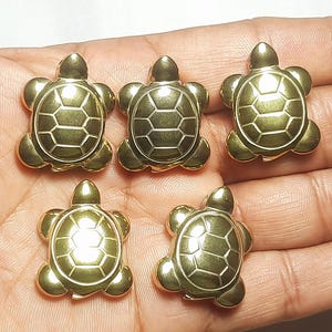 May include: Five gold-coloured turtle charms. Each charm features a detailed turtle shell design, with the head, legs, and shell clearly defined. The charms are arranged on a light-coloured surface, showcasing their metallic sheen.