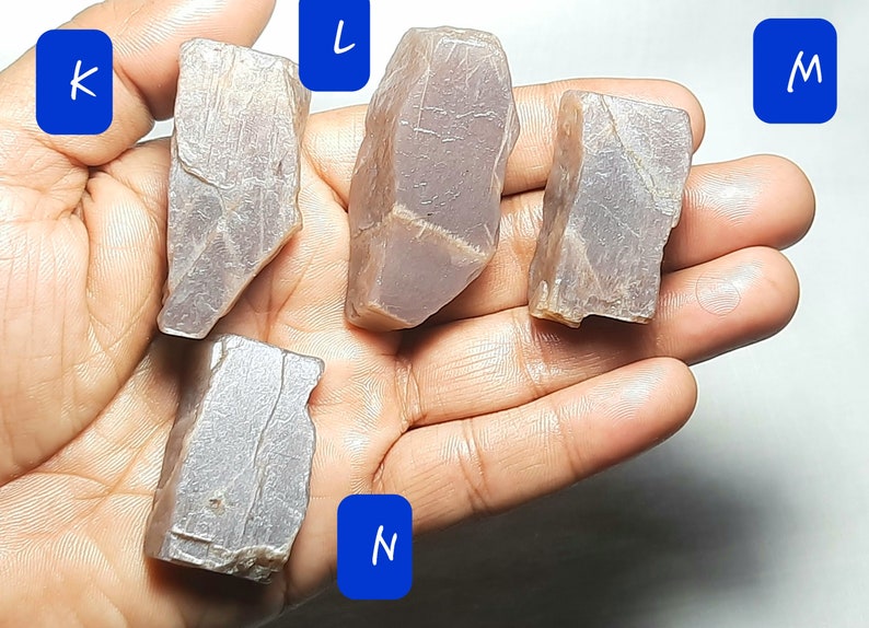 Puede incluir: Cuatro piezas de piedra de sol en bruto, de color p&uacute;rpura claro. Las piedras son de varias formas y tama&ntilde;os. Las piedras est&aacute;n etiquetadas como K, L, M y N.