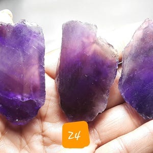 K&ouml;nnte beinhalten: Drei rohe Amethyst-Kristallproben mit verschiedenen Lila-T&ouml;nen und durchscheinenden Bereichen. Die Kristalle werden in einer Hand gehalten, mit orangefarbenen Etiketten mit der Aufschrift Z3, Z4 und Z5.