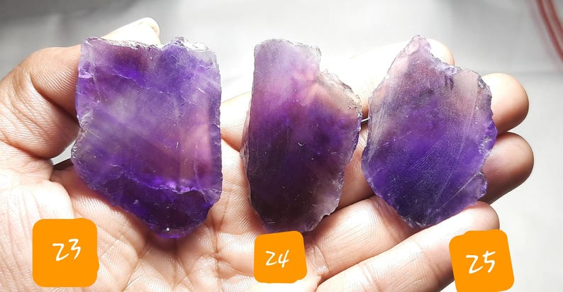 K&ouml;nnte beinhalten: Drei rohe Amethyst-Kristallproben mit verschiedenen Lila-T&ouml;nen und durchscheinenden Bereichen. Die Kristalle werden in einer Hand gehalten, mit orangefarbenen Etiketten mit der Aufschrift Z3, Z4 und Z5.