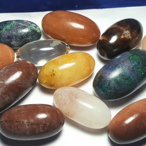Può includere: Una collezione di dieci pietre lisce di forma ovale in vari colori, tra cui marrone, rosso, giallo, verde e bianco. Le pietre sono disposte su una superficie bianca.