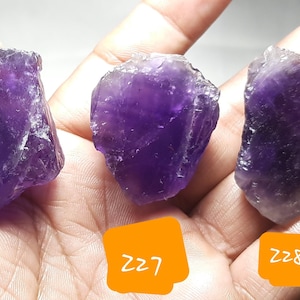 K&ouml;nnte beinhalten: Drei rohe Amethystkristalle in verschiedenen Lilat&ouml;nen werden gezeigt. Jeder Stein hat eine raue, nat&uuml;rliche Textur und wird in einer Hand gehalten. Orangefarbene Etiketten mit den Nummern 226, 227 und 228 sind sichtbar.