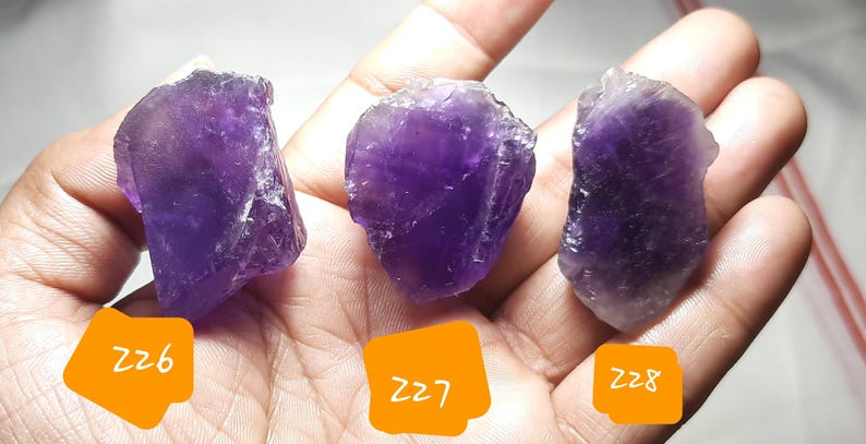K&ouml;nnte beinhalten: Drei rohe Amethystkristalle in verschiedenen Lilat&ouml;nen werden gezeigt. Jeder Stein hat eine raue, nat&uuml;rliche Textur und wird in einer Hand gehalten. Orangefarbene Etiketten mit den Nummern 226, 227 und 228 sind sichtbar.