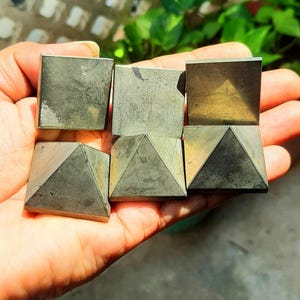 May include: Six pyrite cubes and pyramids are held in a hand. The cubes and pyramids have a metallic, grey-gold colour with a slightly textured surface. The objects are arranged in a grid-like pattern, with three cubes on top and three pyramids below.