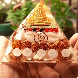 May include: A clear pyramid with a gold-colored miniature temple inside. The pyramid is filled with brown seeds, white seashells, and a copper wire spiral.