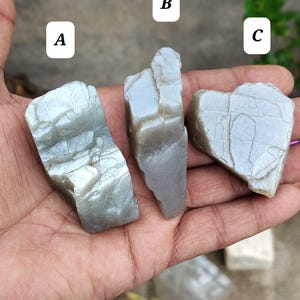 May include: Three raw, light gray, translucent mineral stones labeled A, B, and C. Each stone features a jagged, irregular shape with visible surface cracks and a smooth, slightly reflective texture. The stones are displayed in a palm.