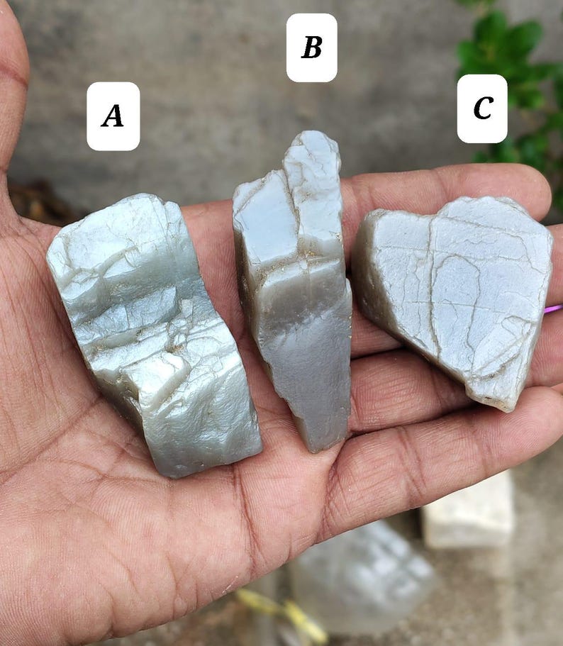 May include: Three raw, light gray, translucent mineral stones labeled A, B, and C. Each stone features a jagged, irregular shape with visible surface cracks and a smooth, slightly reflective texture. The stones are displayed in a palm.
