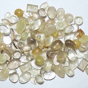 May include: A collection of translucent, faceted, and polished rutilated quartz gemstones in various shapes and sizes. The stones have a light golden color with visible inclusions of golden rutile needles.