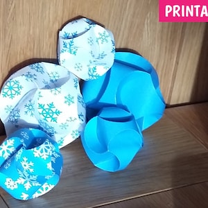 Puede incluir: Cuatro adornos de papel con un patr&oacute;n de copo de nieve. Dos son azules y dos son blancos con copos de nieve azules. Los adornos est&aacute;n sobre una superficie de madera. PRINTABLE est&aacute; escrito en rosa en la parte superior de la imagen.