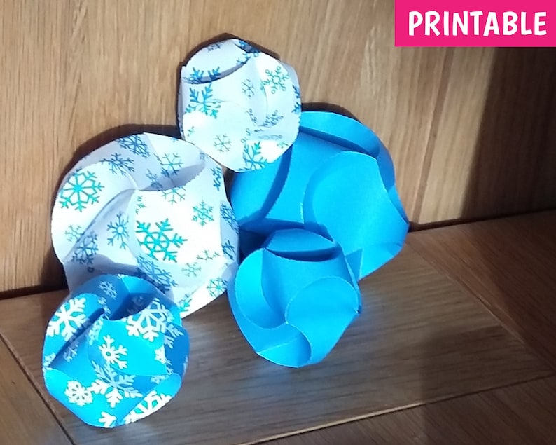 Puede incluir: Cuatro adornos de papel con un patr&oacute;n de copo de nieve. Dos son azules y dos son blancos con copos de nieve azules. Los adornos est&aacute;n sobre una superficie de madera. PRINTABLE est&aacute; escrito en rosa en la parte superior de la imagen.