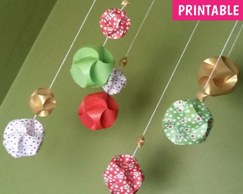 Puede incluir: Adornos de papel imprimibles en varios colores y patrones, incluyendo rojo, verde, dorado y blanco. Los adornos cuelgan de cuerdas y presentan diferentes dise&ntilde;os, como estrellas, lunares y formas geom&eacute;tricas. La palabra "IMPRIMIBLE" es visible en la esquina superior derecha de la imagen.