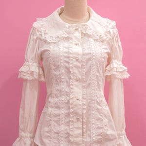 Op de afbeelding: Witte blouse met lange mouwen, een gerimpelde kraag en manchetten, op een mannequin. De blouse heeft delicate kanten details, parelmoer knopen en verticale kanten afwerking. De achtergrond is effen roze.