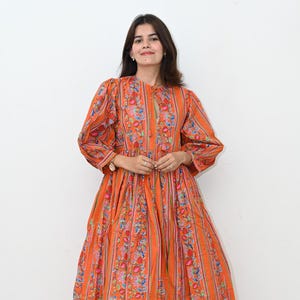 May include: A long orange dress with a floral and striped design. The dress features long sleeves and a keyhole neckline. The model is wearing white sandals. The dress is a striking and colourful garment.