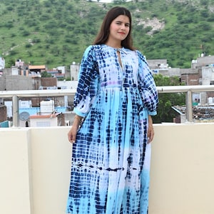 Könnte beinhalten: Ein langes, fließendes Kleid mit einem Batikmuster in Blau- und Weißtönen. Das Kleid hat einen Schlüssellochausschnitt und lange, puffärmelige Ärmel. Das Model trägt weiße Sandalen. Der Hintergrund umfasst eine Stadtlandschaft und grüne Hügel.