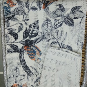 May include: A white kantha quilt with a black and blue floral pattern. The quilt is folded in half and is sitting on a woven bamboo surface.