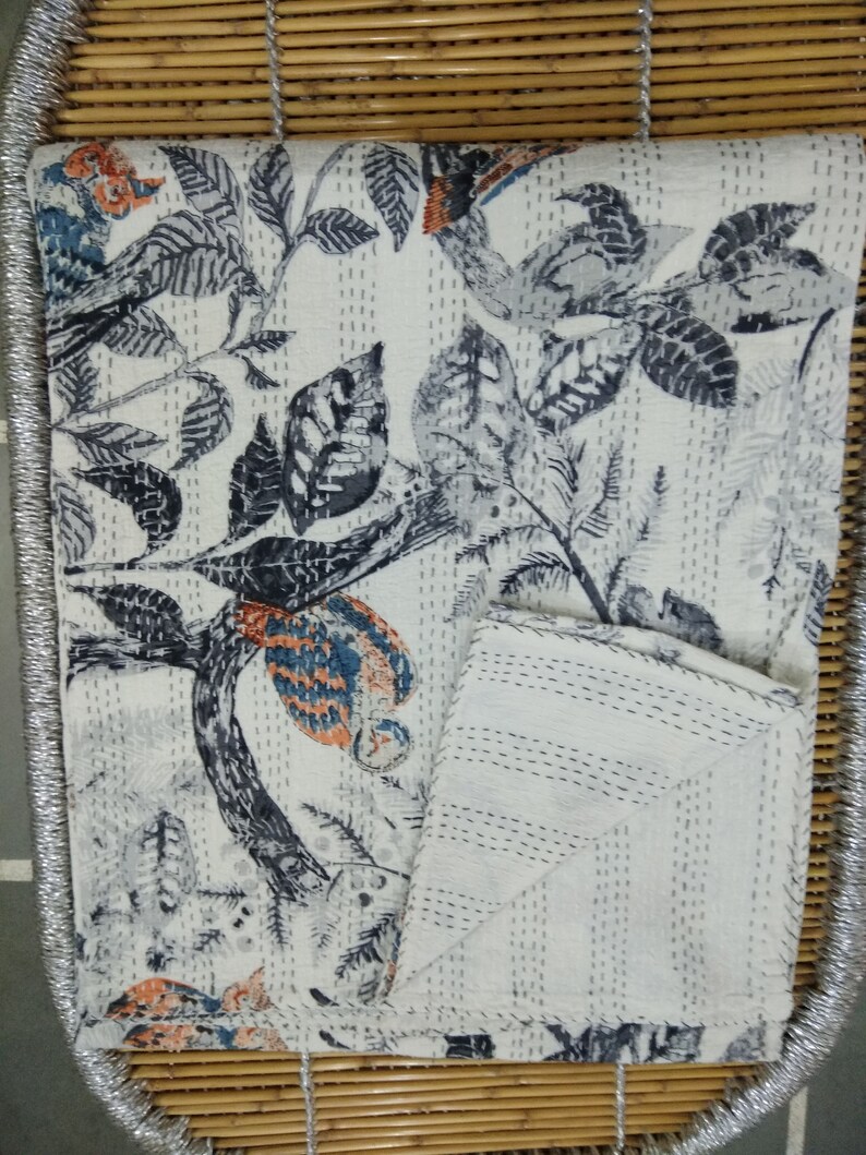 May include: A white kantha quilt with a black and blue floral pattern. The quilt is folded in half and is sitting on a woven bamboo surface.
