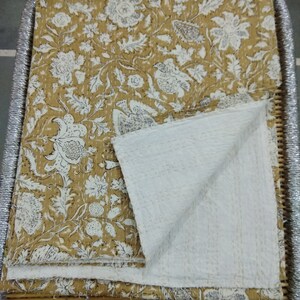 May include: A kantha quilt with a floral pattern in shades of white and brown on a mustard yellow background. The quilt is folded in half and is lying on a woven bamboo tray.