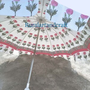 Op de afbeelding: Een witte en roze parasol met een bloemmotief, een rode rand en een zilveren handvat. De parasol is open en het handvat wijst naar beneden. De parasol staat op een grijs betonnen oppervlak. De tekst "Ruralartandcraft" is zichtbaar op de parasol.