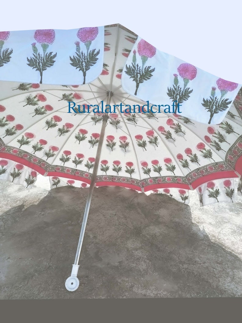 Op de afbeelding: Een witte en roze parasol met een bloemmotief, een rode rand en een zilveren handvat. De parasol is open en het handvat wijst naar beneden. De parasol staat op een grijs betonnen oppervlak. De tekst "Ruralartandcraft" is zichtbaar op de parasol.