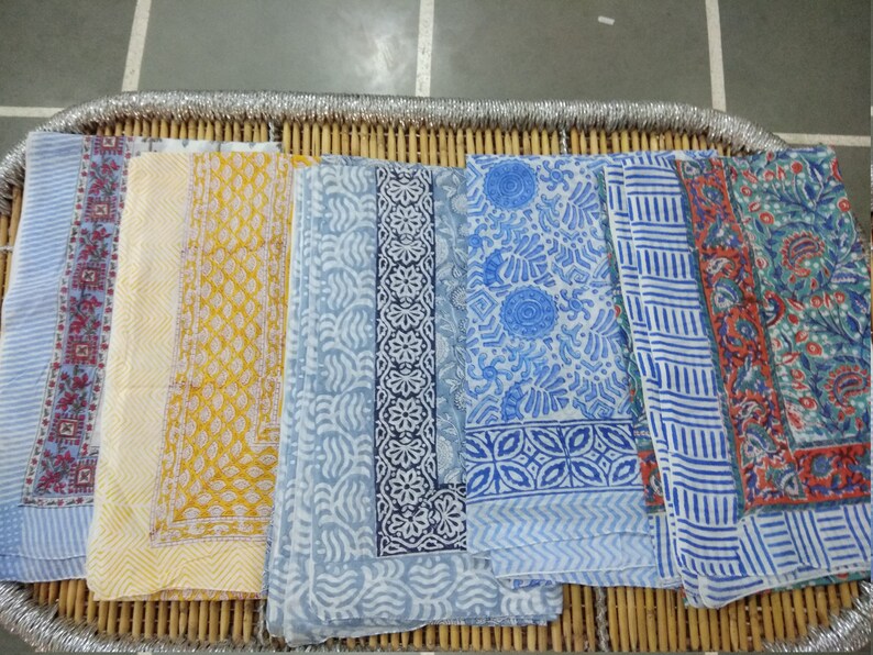 May include: Five different patterned fabric squares in shades of blue, yellow, and red. The fabric squares are arranged in a row on a woven bamboo surface.