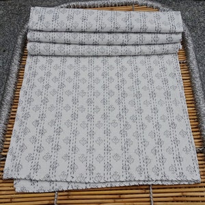 May include: A stack of white cotton fabric with a repeating pattern of small black flowers and lines. The fabric is folded in thirds and has a slightly rough texture.