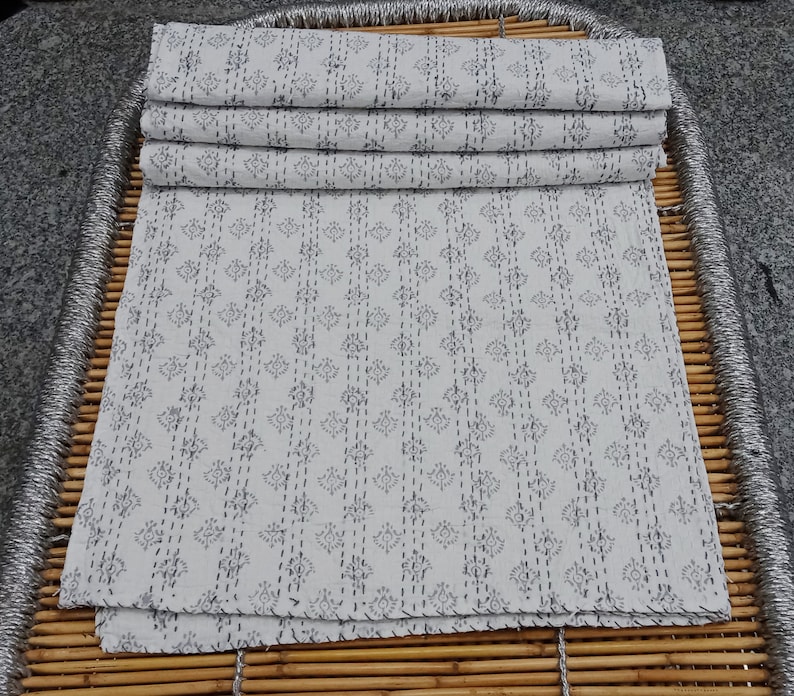 May include: A stack of white cotton fabric with a repeating pattern of small black flowers and lines. The fabric is folded in thirds and has a slightly rough texture.