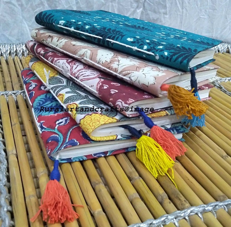 May include: A stack of four handmade journals with colorful floral patterned covers and tassels. The journals are on a woven bamboo surface.