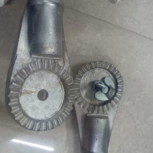 Op de afbeelding: Twee zilveren metalen onderdelen met een tandwielachtig ontwerp. De onderdelen zijn verbonden door een metalen staaf. De onderdelen worden gebruikt om de hoogte van een stoel of tafel aan te passen.