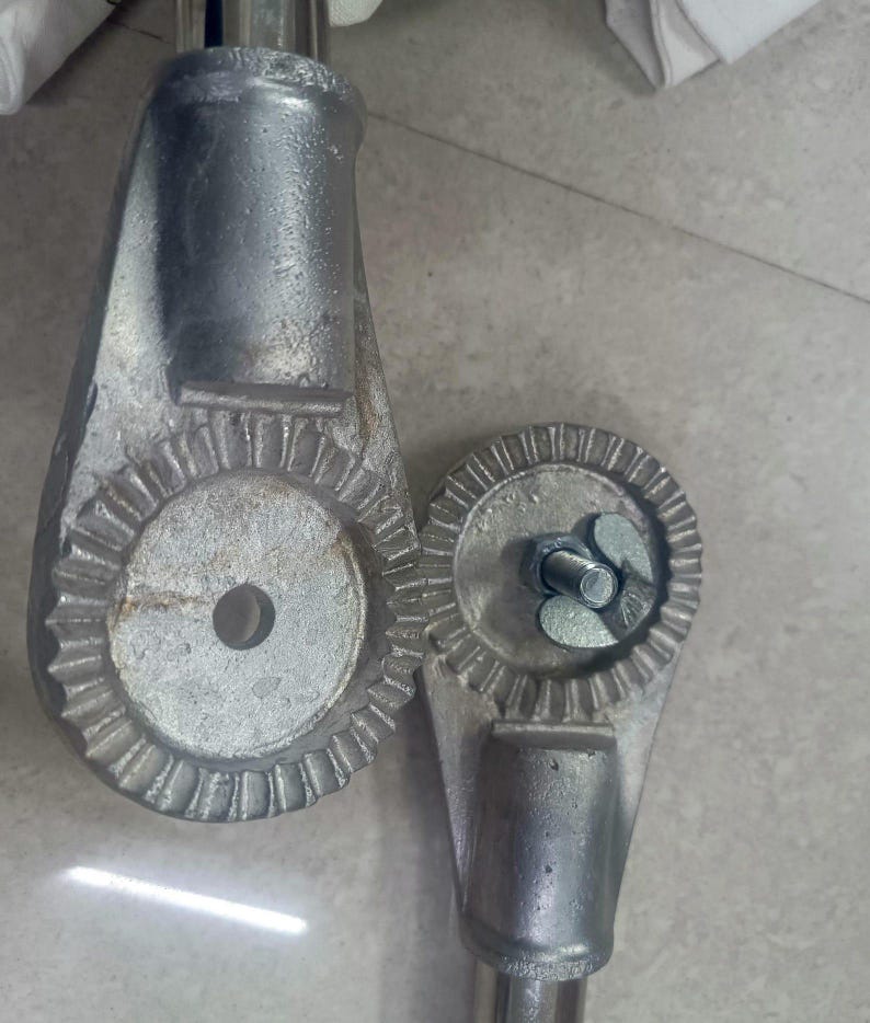 Op de afbeelding: Twee zilveren metalen onderdelen met een tandwielachtig ontwerp. De onderdelen zijn verbonden door een metalen staaf. De onderdelen worden gebruikt om de hoogte van een stoel of tafel aan te passen.