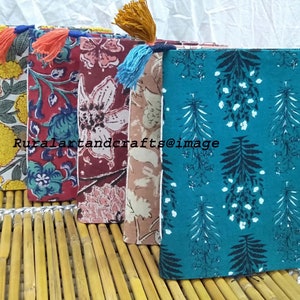 May include: Four handmade fabric-covered journals with colorful floral and leaf patterns. Each journal has a tassel bookmark and is bound with a white stitch. The journals are arranged in a row on a bamboo surface.