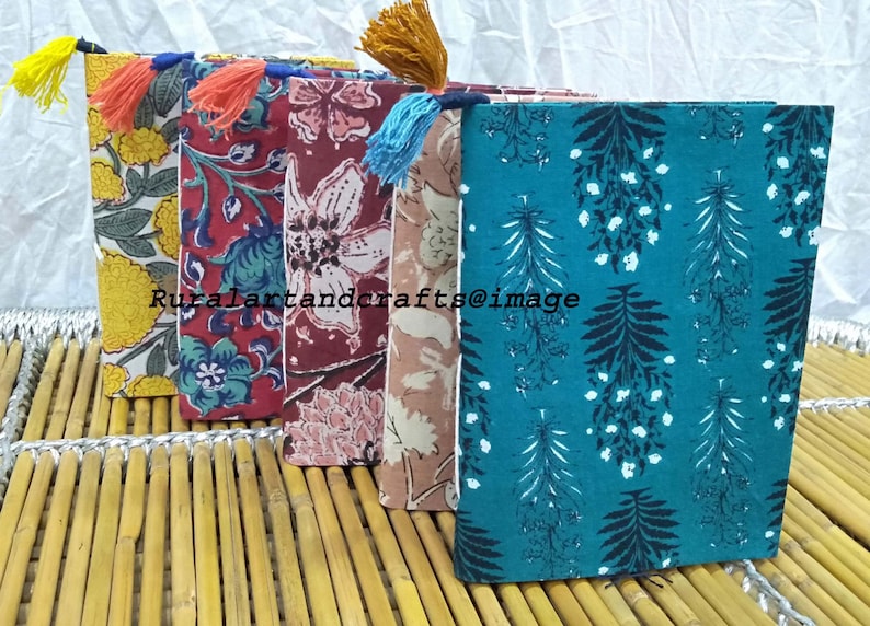 May include: Four handmade fabric-covered journals with colorful floral and leaf patterns. Each journal has a tassel bookmark and is bound with a white stitch. The journals are arranged in a row on a bamboo surface.