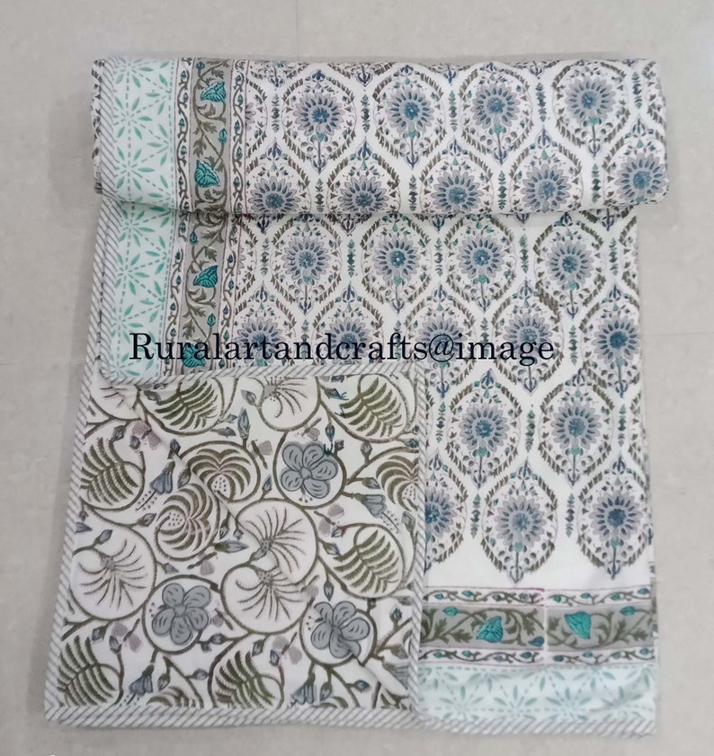 May include: A white cotton quilt with a floral pattern in shades of blue, green, and gray. The quilt is folded in half and has a border with a repeating pattern.