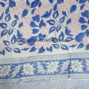 Op de afbeelding: Witte katoenen stof met een blauwe en roze bloemenprint. De stof heeft een rand met een blauw en wit bloemenpatroon en een zigzagpatroon.