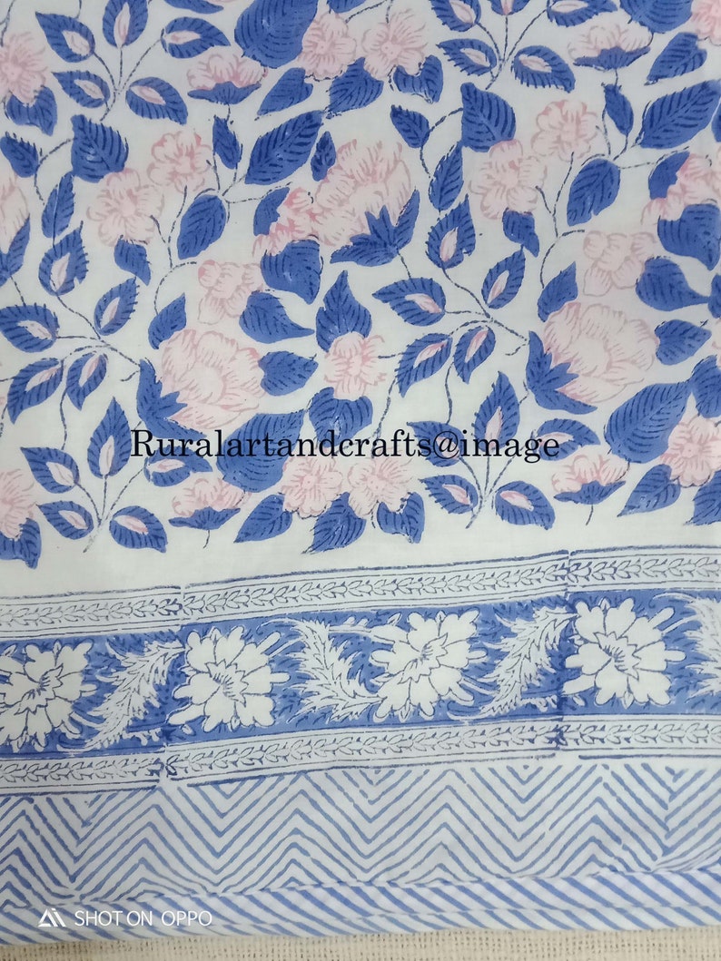 Op de afbeelding: Witte katoenen stof met een blauwe en roze bloemenprint. De stof heeft een rand met een blauw en wit bloemenpatroon en een zigzagpatroon.
