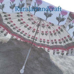 Op de afbeelding: Een witte en roze bloemenprint paraplu met een zilveren handvat. De paraplu is open en de stof is bedrukt met roze bloemen en groene bladeren. De tekst "Ruralartandcraft" is op de stof gedrukt.