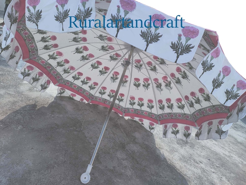 Op de afbeelding: Een witte en roze bloemenprint paraplu met een zilveren handvat. De paraplu is open en de stof is bedrukt met roze bloemen en groene bladeren. De tekst "Ruralartandcraft" is op de stof gedrukt.