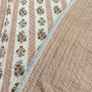 May include: A quilted blanket with a floral pattern and striped design. The blanket features alternating vertical stripes in brown and white, with floral motifs in brown and grey. The edge has a blue zigzag pattern.