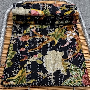 May include: A black kantha quilt with a floral pattern in shades of green, pink, and white. The quilt is folded and displayed on a woven bamboo tray.
