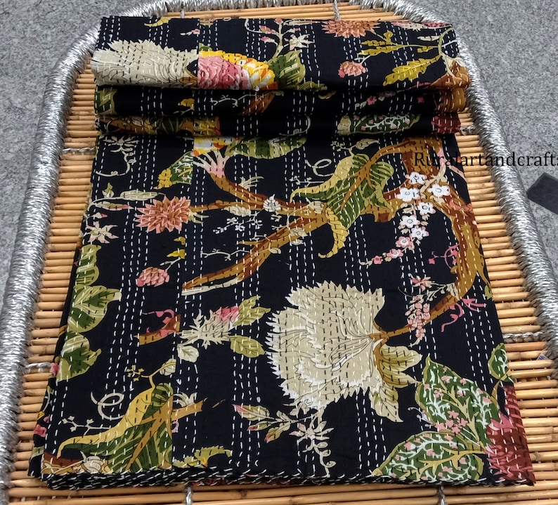 May include: A black kantha quilt with a floral pattern in shades of green, pink, and white. The quilt is folded and displayed on a woven bamboo tray.