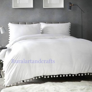 May include: A white bedding set with a duvet cover and two pillowcases. The set is trimmed with small white pom-poms along the edges. The bedding is displayed on a bed with a gray headboard and a white rug.