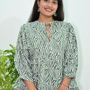 May include: A long-sleeved blouse featuring an olive green and white zebra stripe pattern, a round neckline with a tie closure, and a gathered peplum waist. The garment has loose, voluminous sleeves that end at the elbow with a straight hemline.