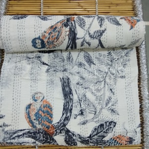 May include: A white kantha quilt with a repeating pattern of birds and trees. The quilt is folded in half and is lying on a woven bamboo mat.